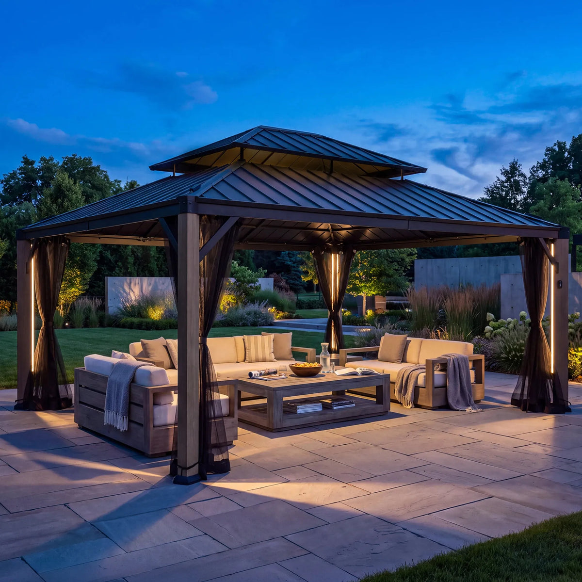 A large outdoor double-tiered gazebo with dark metal roofing and draped netting, furnished with a beige sectional sofa, coffee table, and string lights on a stone patio at dusk.