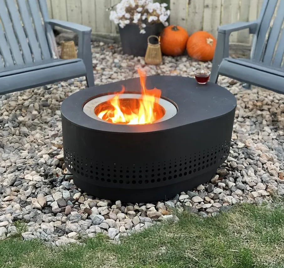 Black fire pit with flames in a garden setting with chairs and pumpkins.