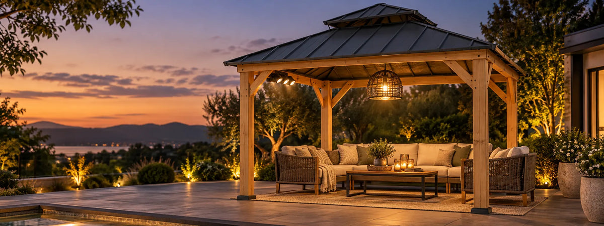 A wooden outdoor patio gazebo with a metal roof, furnished with a sofa, chairs, and a coffee table under warm lights, set on a stone patio overlooking a scenic sunset view with mountains and water in the background.