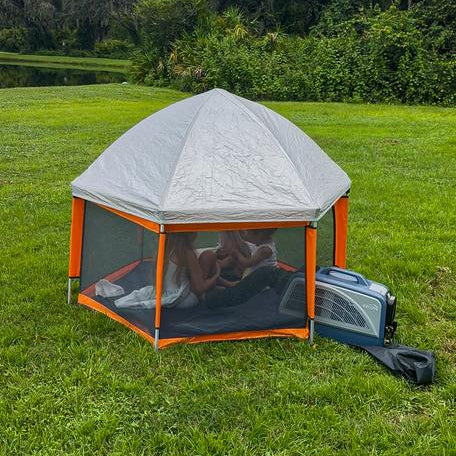 A small pop-up canopy tent with orange trim and mesh sides is set up on a grassy lawn outdoors. Two children are visible inside the tent, and a blue portable air conditioner sits beside it on the grass, with trees and water in the background.