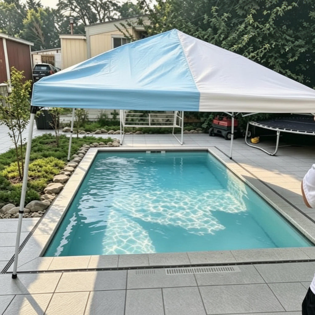 A rectangular swimming pool with turquoise water in a backyard setting, shaded by a light blue and white pop-up canopy tent. The pool is surrounded by a gray tiled deck, with trees, houses, and a trampoline visible in the background.
